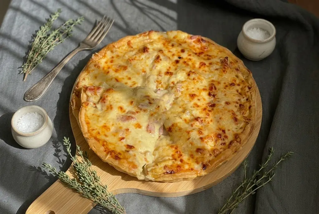 Une main gantée enfourne la tarte tartiflette recouverte de fromage râpé dans un four chaud et illuminé pour la cuisson finale et le gratinage doré.