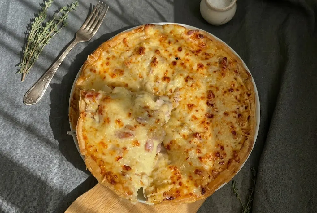 Une main gantée enfourne la tarte tartiflette recouverte de fromage râpé dans un four chaud et illuminé pour la cuisson finale et le gratinage doré.
